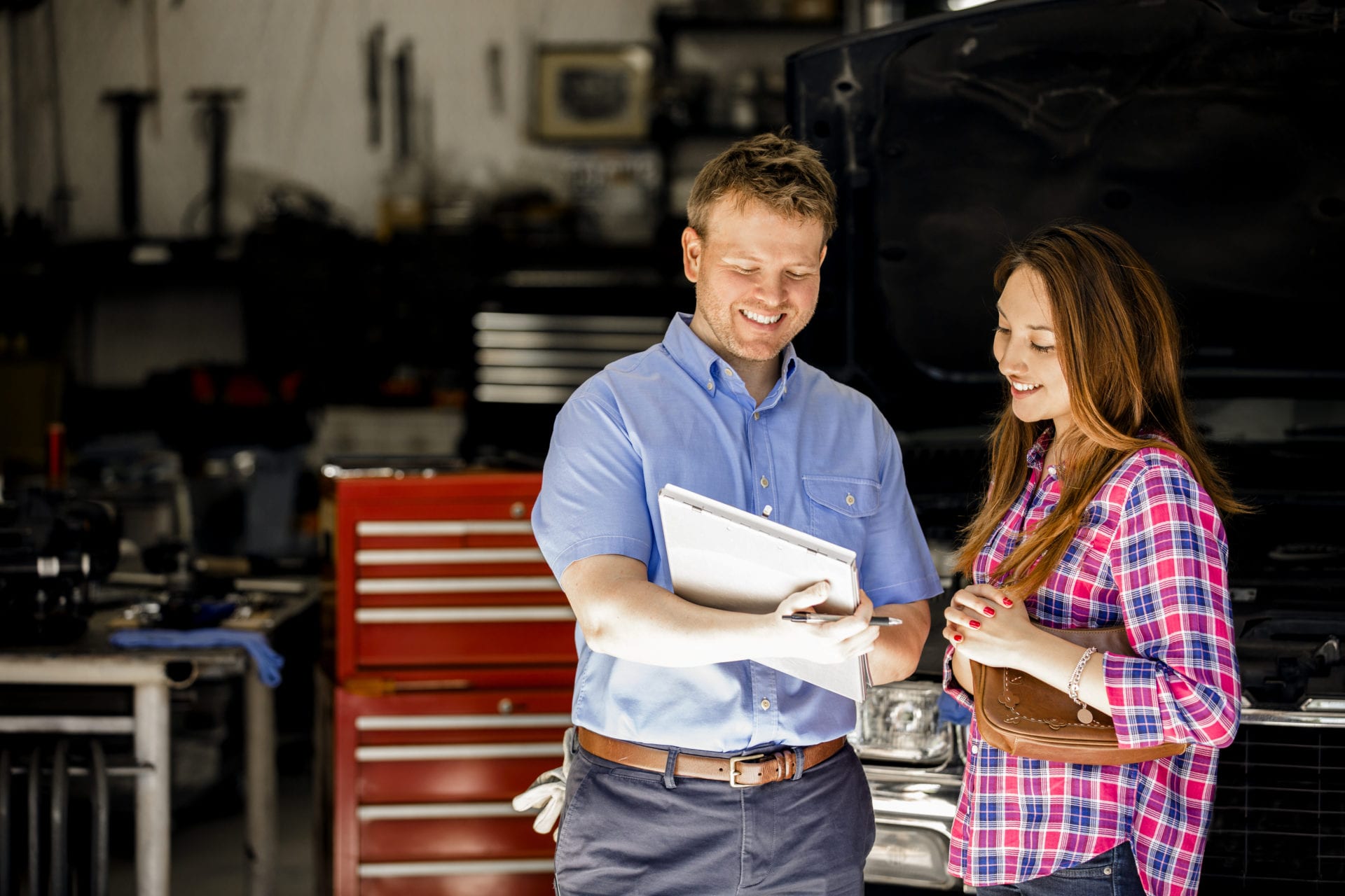 Happy customer!  A smiling Latin descent woman discusses automobile repair invoice with an auto mechanic in a repair shop.  She is discussing the vehicle's repairs with the mechanic, who is explaining her service.  Toolbox, workshop background. Unidentifiable, SUV-style vehicle.